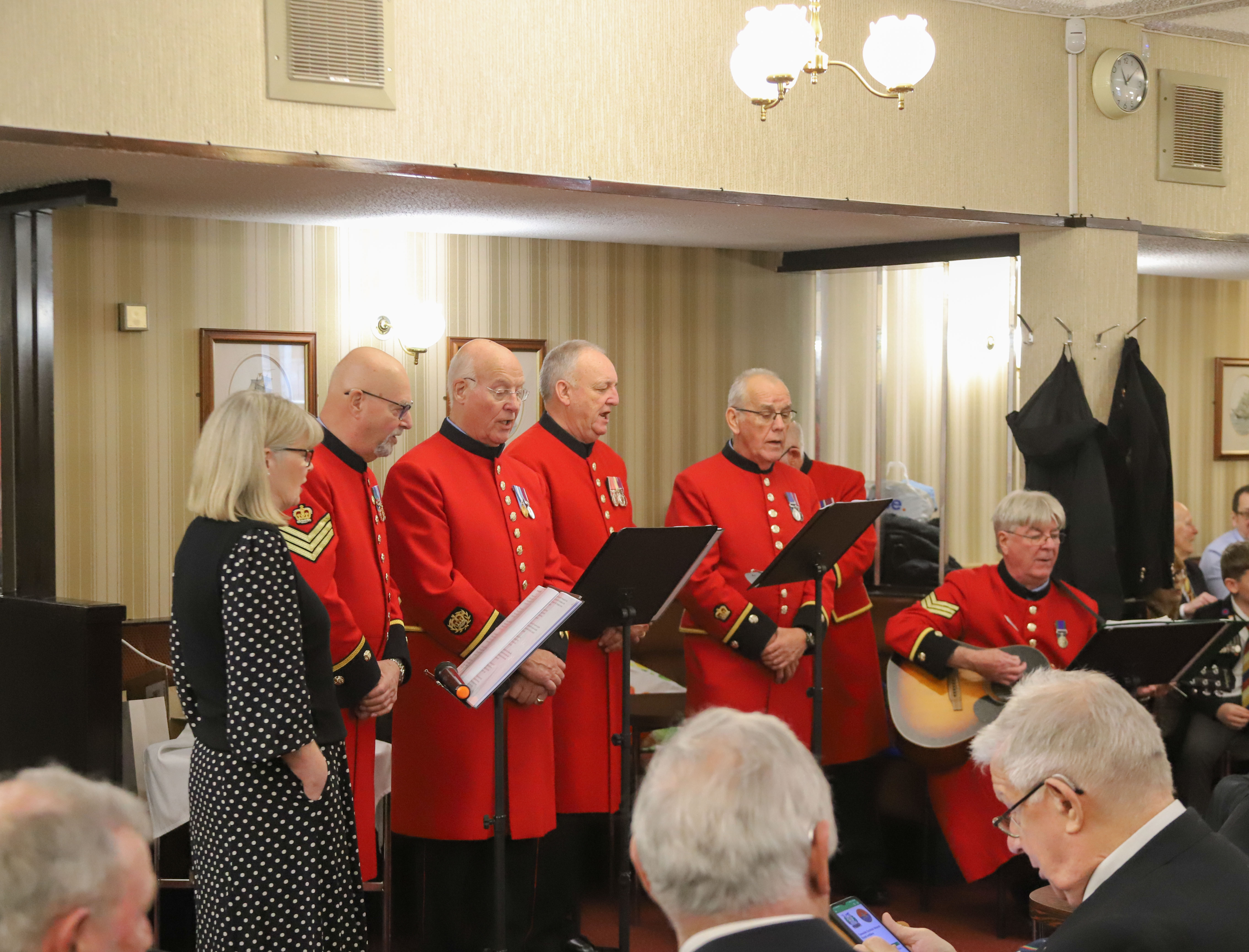 Group of Chelsea Pensioners in scarlet uniform stood on a small stage singing