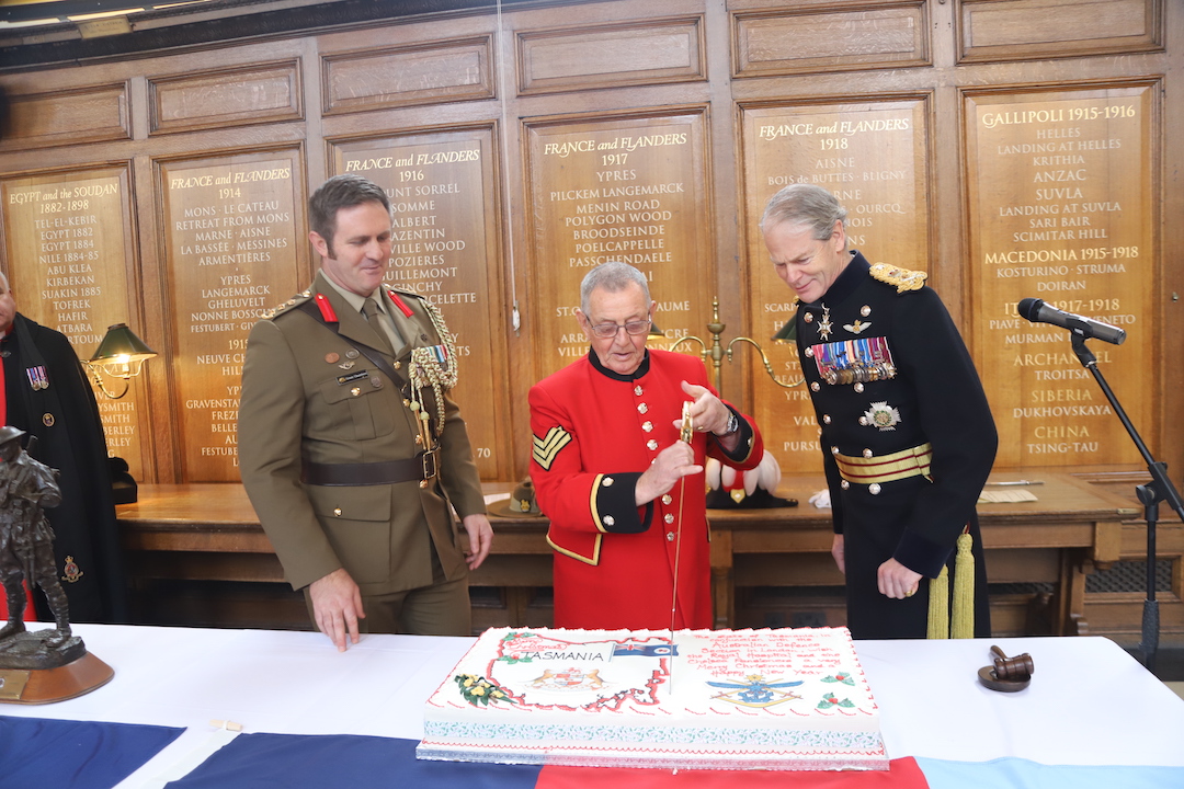 Chelsea Pensioner Yorkie cuts a cake with a ceremonial sword and the Governor and Col clapping stood either side