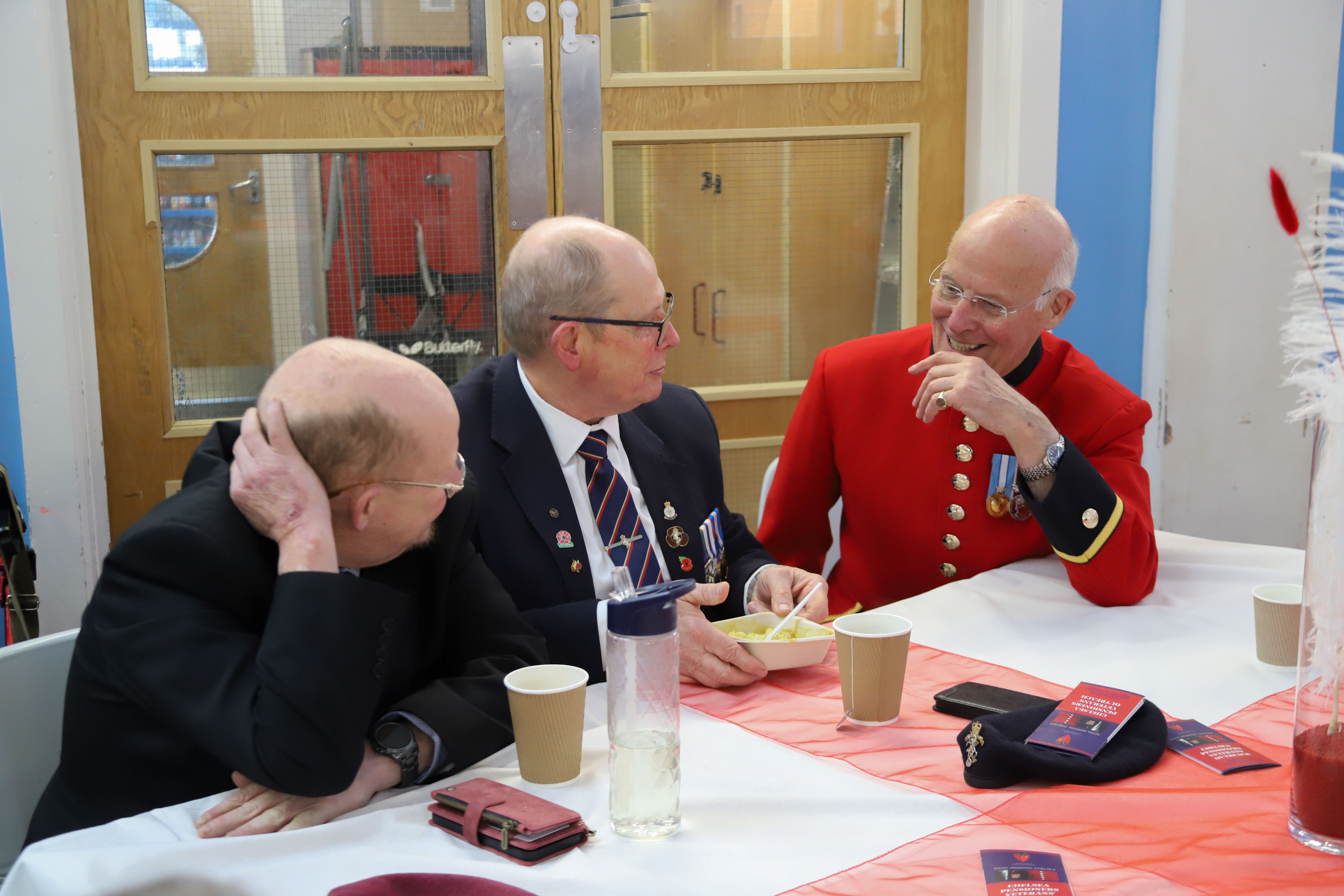 Chelsea Pensioner in scarlet uniform sat with two Veterans talking and smiling