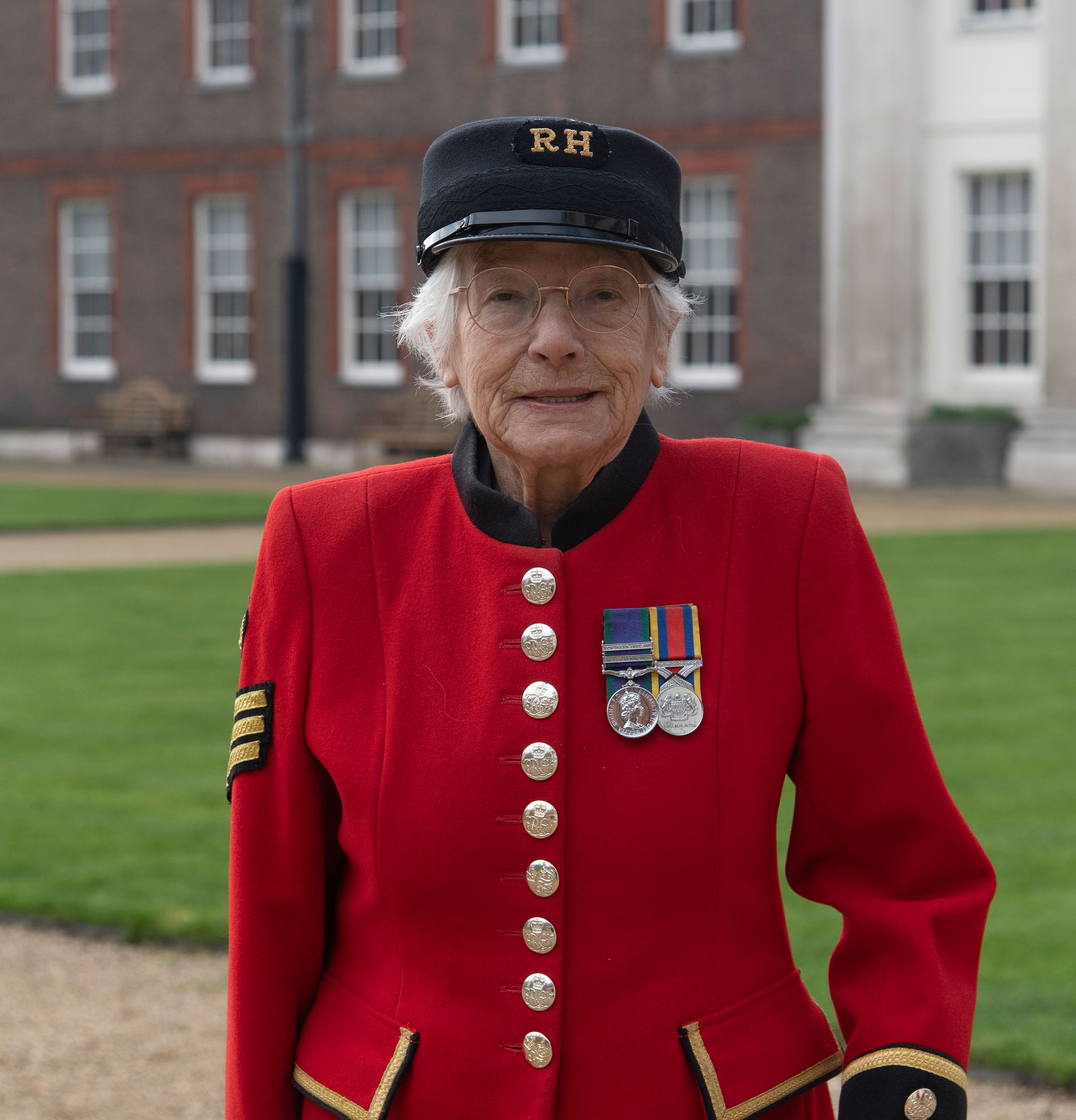 Female Chelsea Pensioner, Marjorie Cole, in Scarlet uniform and shako smiles at the camera