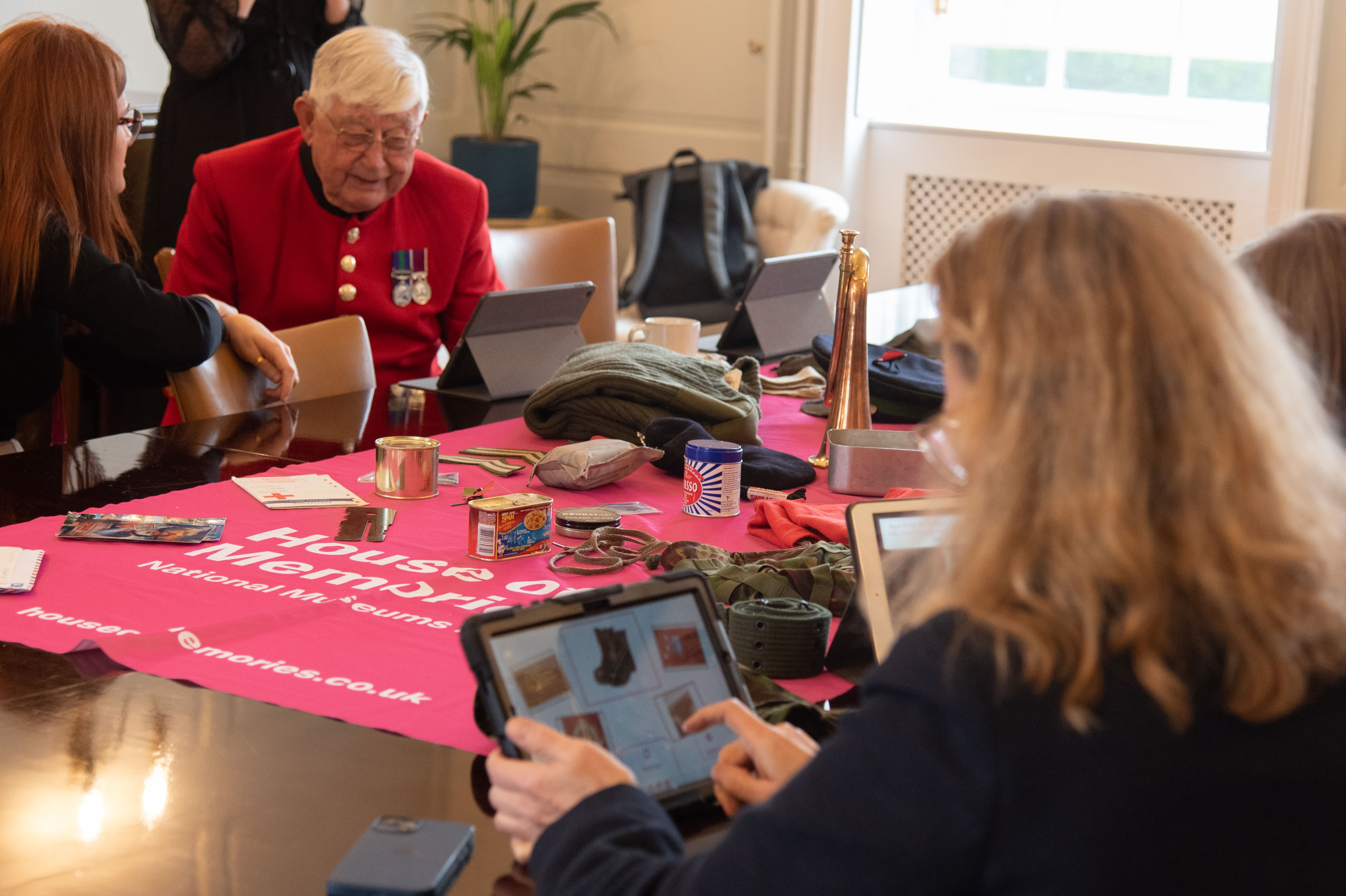 People sat round a table looking at iPads. A pink cloth is in the middle of the table with the House of Memories logo on and objects from the My House of Memories app