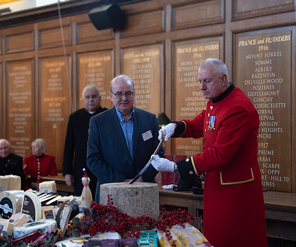 Chelsea Pensioner in scarlet uniform holding a sword to cut a 25kg Montgomery cheddar cheese